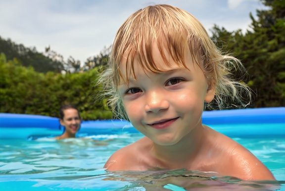 Ein blonder Junge hat Spaß in einen Swimming Pool, im Hintergrund ist eine junge Frau zu sehen